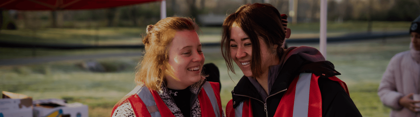 Two girls laughing with each other outside during a conversation wearing red safety vests.